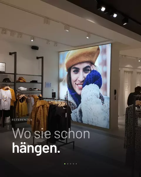 Feed-Kachel: In einem Modegeschäft leuchtet eine Wand mit dem Porträt einer Frau mit Mütze, darüber „Wo sie schon hängen."