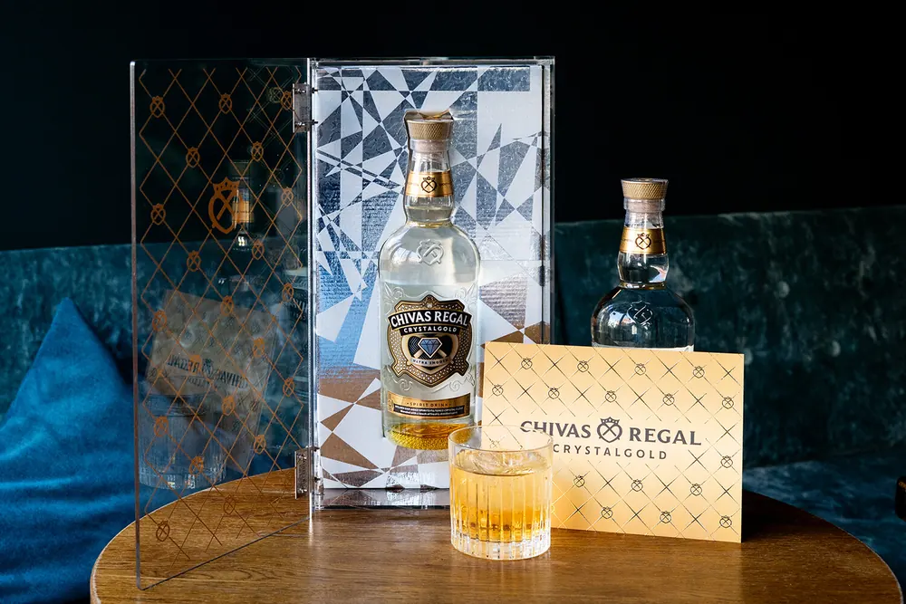 Aufgeklappte Acrylglas-Box mit goldenem Rautenmuster, darin eine Flasche Chivas Regal Crystalgold, davor ein gefülltes Glas und eine Karte