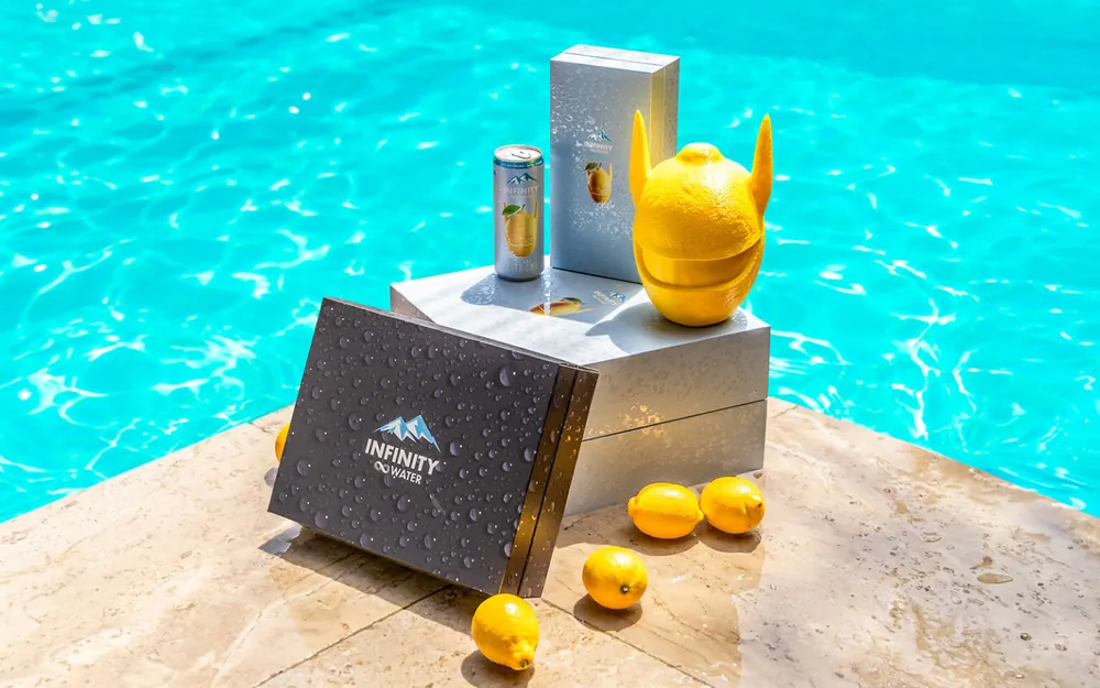 Silberne und schwarze Verpackungen am Rand eines türkisfarbenen Pools, daneben eine Zitrone in Form einer Pandamaske und eine Dose Infinity Water