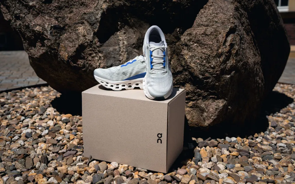 Ein Paar hellgraue On-Laufschuhe steht auf einer schlichten Graupappschachtel vor einem großen Felsen